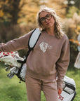 Woman in a pink sweatshirt and pants holding golf clubs with another person in the background.