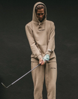 Person wearing a beige tracksuit with 'Golf Shirt' branding, holding a golf club on a dark background
