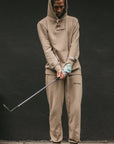 Person wearing a beige hoodie and pants with a brand logo, holding a golf club against a dark background.