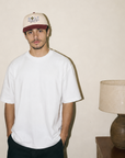 A man wearing a white crewneck t-shirt, dark pants, and a beige baseball cap with a dark red brim, standing in a room with a plain beige wall and a table lamp on a side table.