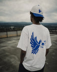 Person wearing a white t-shirt with a blue eagle design on the back, standing on a rooftop with a cityscape in the background.
