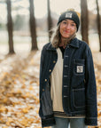 Person wearing a dark jacket and beanie standing in a forest with fallen leaves on the ground.