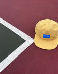 A yellow baseball cap with a blue GOOD > SAFE patch on the front, resting on a red tennis court near the corner line.