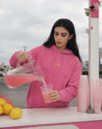 Woman in pink hoodie pouring pink lemonade into a cup at an outdoor stand.