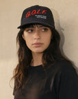 A woman wearing a black baseball cap with red and white embroidered text that reads 'G.O.L.F.