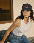 A woman wearing a black baseball cap with red 'GOLF' lettering and a white tank top leans on a bench outdoors next to a building.