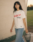 A woman stands outdoors against a light brick wall wearing a white graphic T-shirt with red and black text reading 'GOLF. TO KEEP KIDS OFF DRUGS.