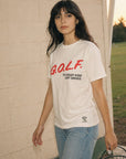 A woman stands outdoors against a light brick wall wearing a white graphic T-shirt with red and black text reading 'GOLF. TO KEEP KIDS OFF DRUGS.