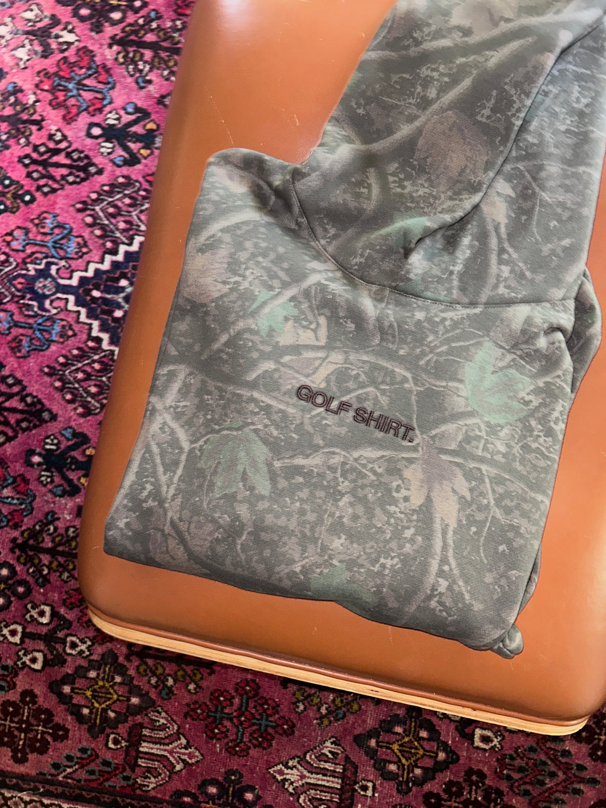 A folded gray camouflage golf shirt with 'GOLF SHIRT' printed on the chest, resting on a tan leather bench over a patterned pink rug.