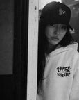 Person wearing a cap and hoodie with text, standing against a wall.