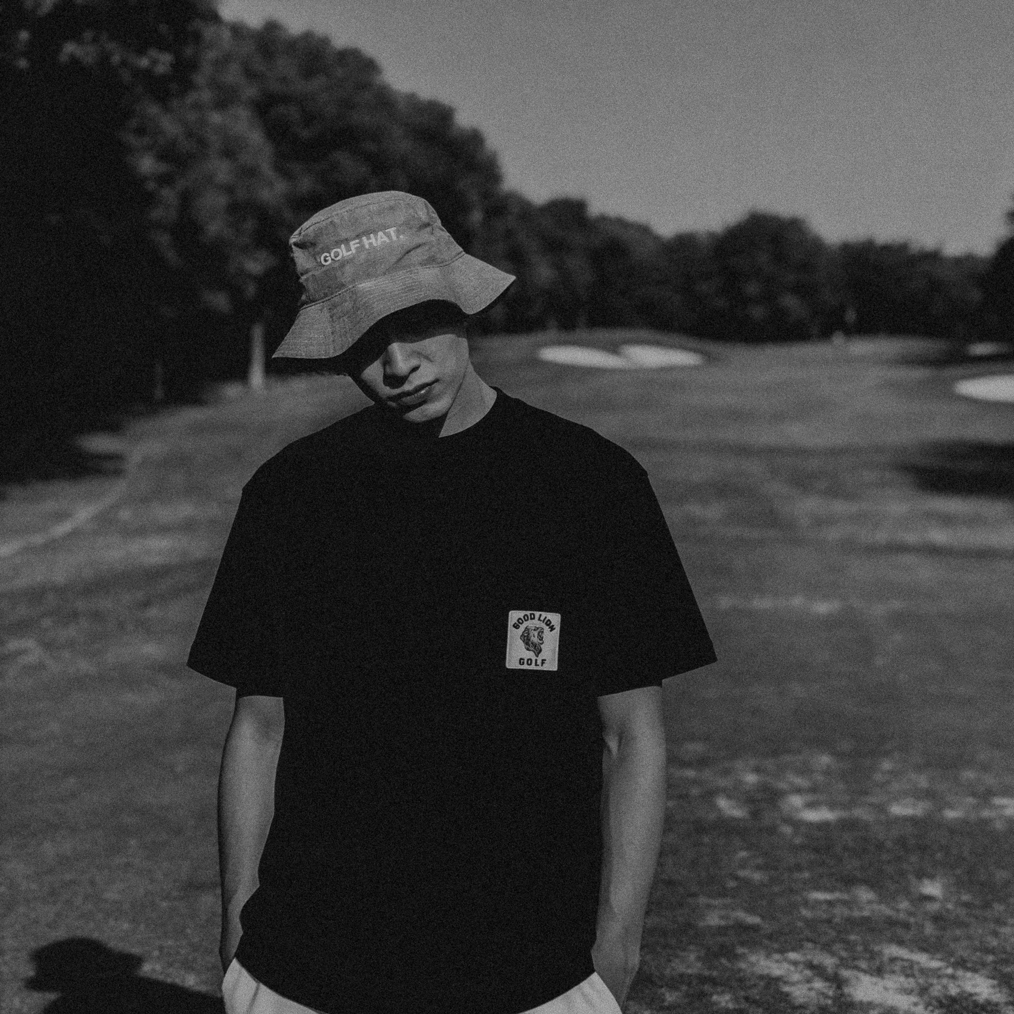 Person wearing a black t-shirt with a logo and a bucket hat on a golf course.