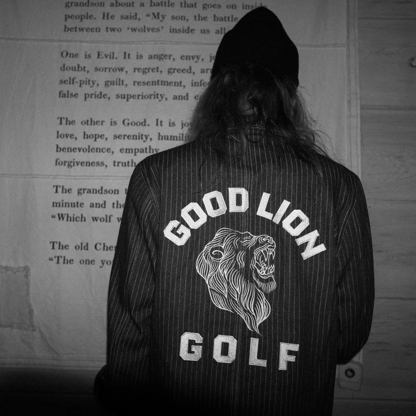Person wearing a jacket with 'Good Lion Golf' logo in front of a wooden wall with text.