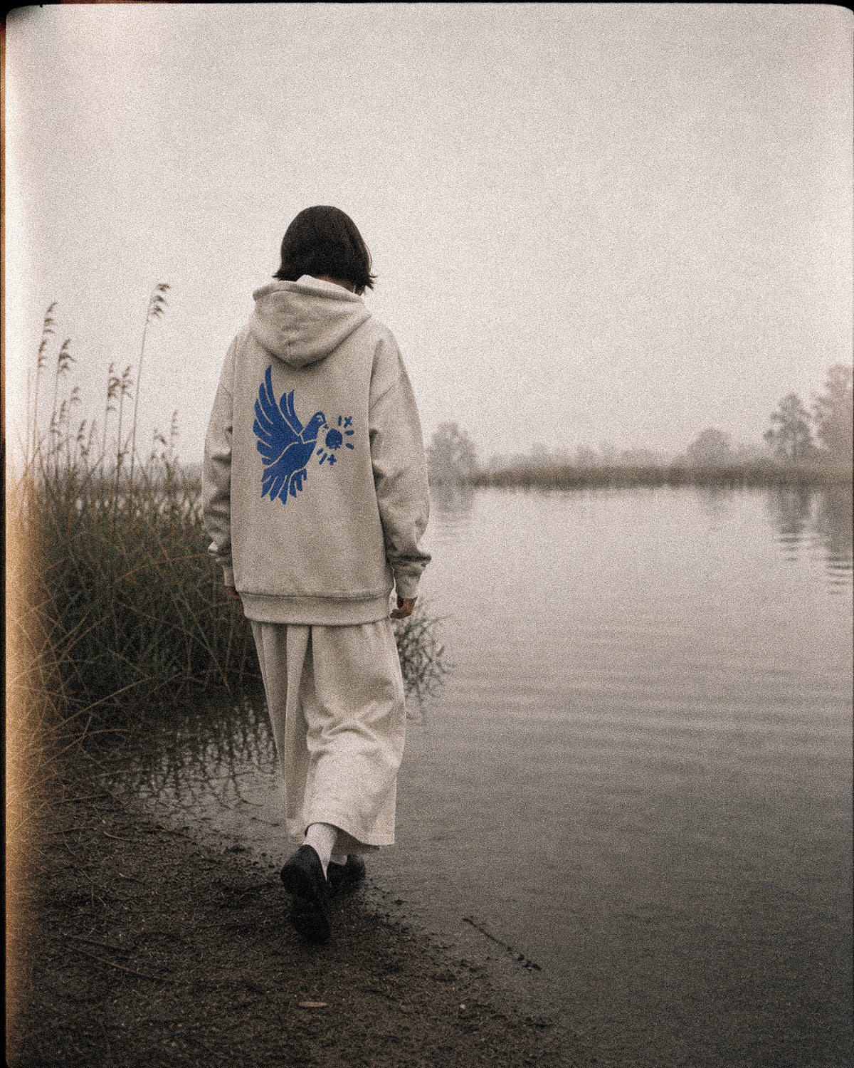 Person wearing a hoodie with a blue dove design walking by a lake.