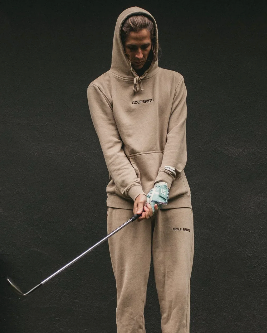 Person wearing a beige tracksuit with 'Golf Shirt' branding, holding a golf club on a dark background
