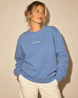 A woman wearing a dusty blue crewneck sweatshirt with 'GOLF SHIRT.' embroidered in white on the chest, paired with cream pants, standing against a textured beige wall.