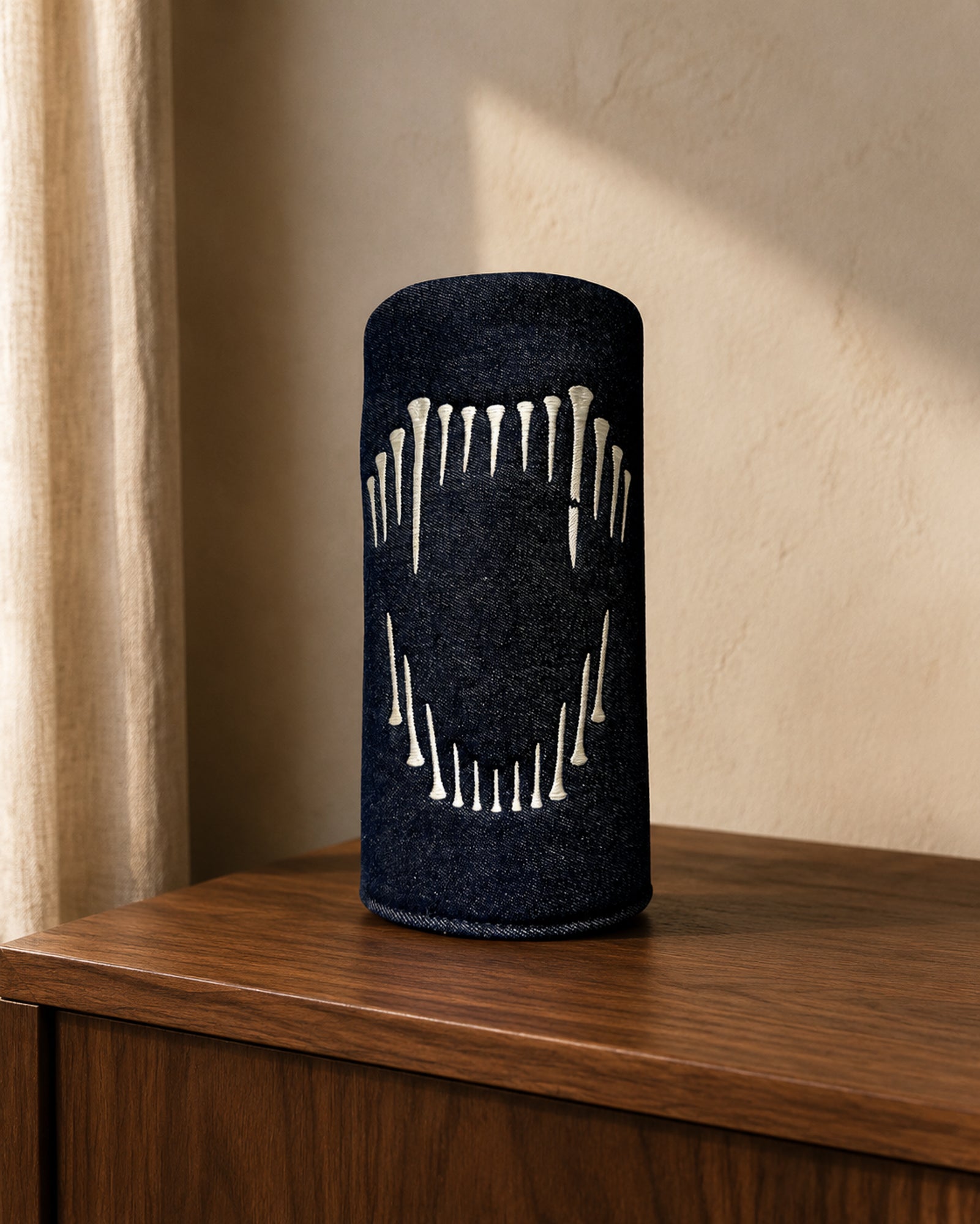 A black cylindrical denim driver cover with a white hand-drawn lion fang design, standing on a wooden cabinet against a textured beige wall.
