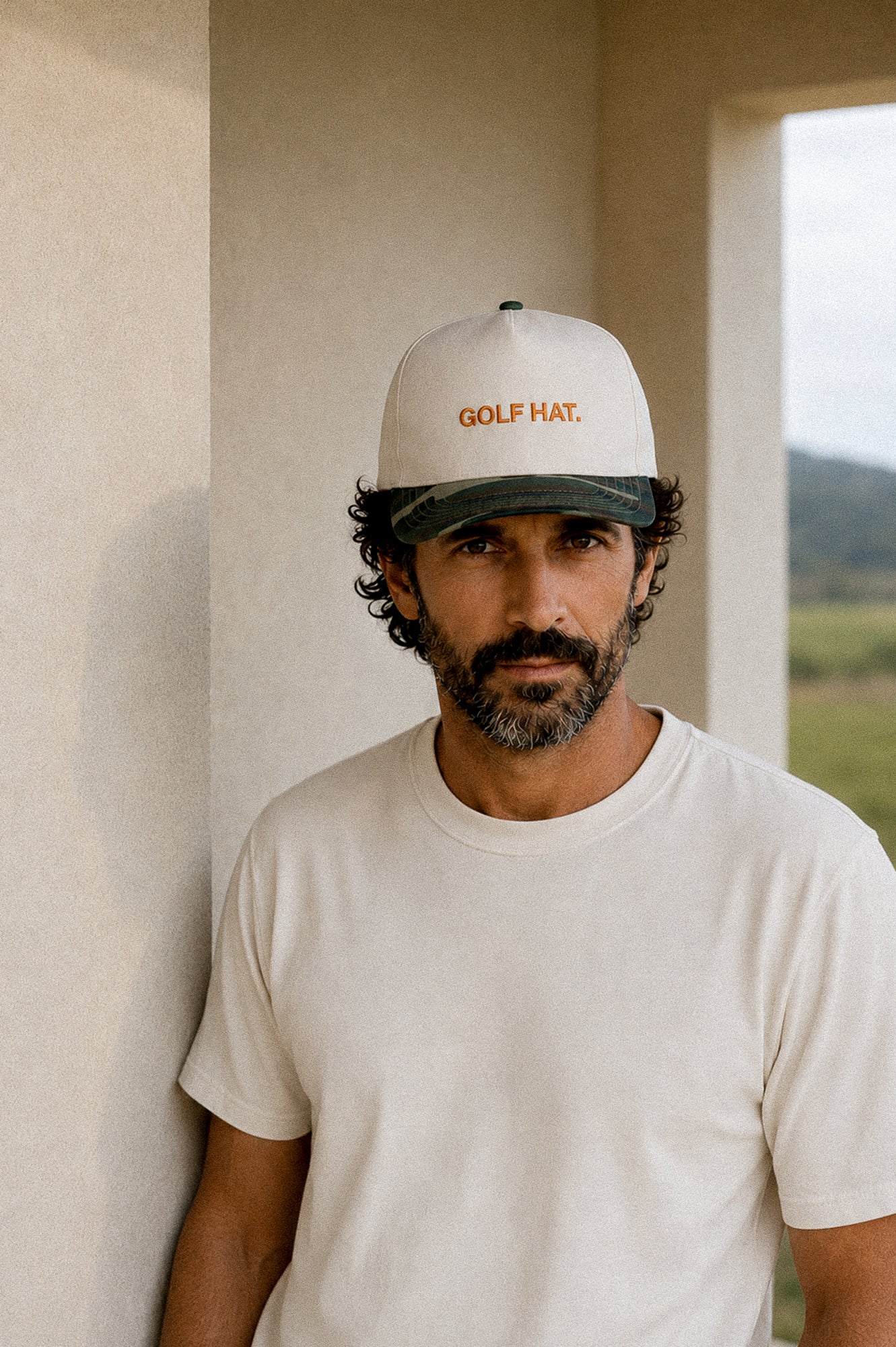 A man wearing a white trucker-style baseball cap with 'GOLF MAT.' embroidered in orange on the front, paired with a white t-shirt, standing in a neutral-toned indoor setting.