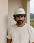 A man wearing a white trucker-style baseball cap with 'GOLF MAT.' embroidered in orange on the front, paired with a white t-shirt, standing in a neutral-toned indoor setting.