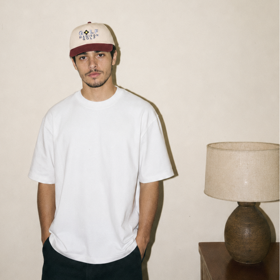 A man wearing a white crewneck t-shirt, dark pants, and a beige baseball cap with a dark red brim, standing in a room with a plain beige wall and a table lamp on a side table.