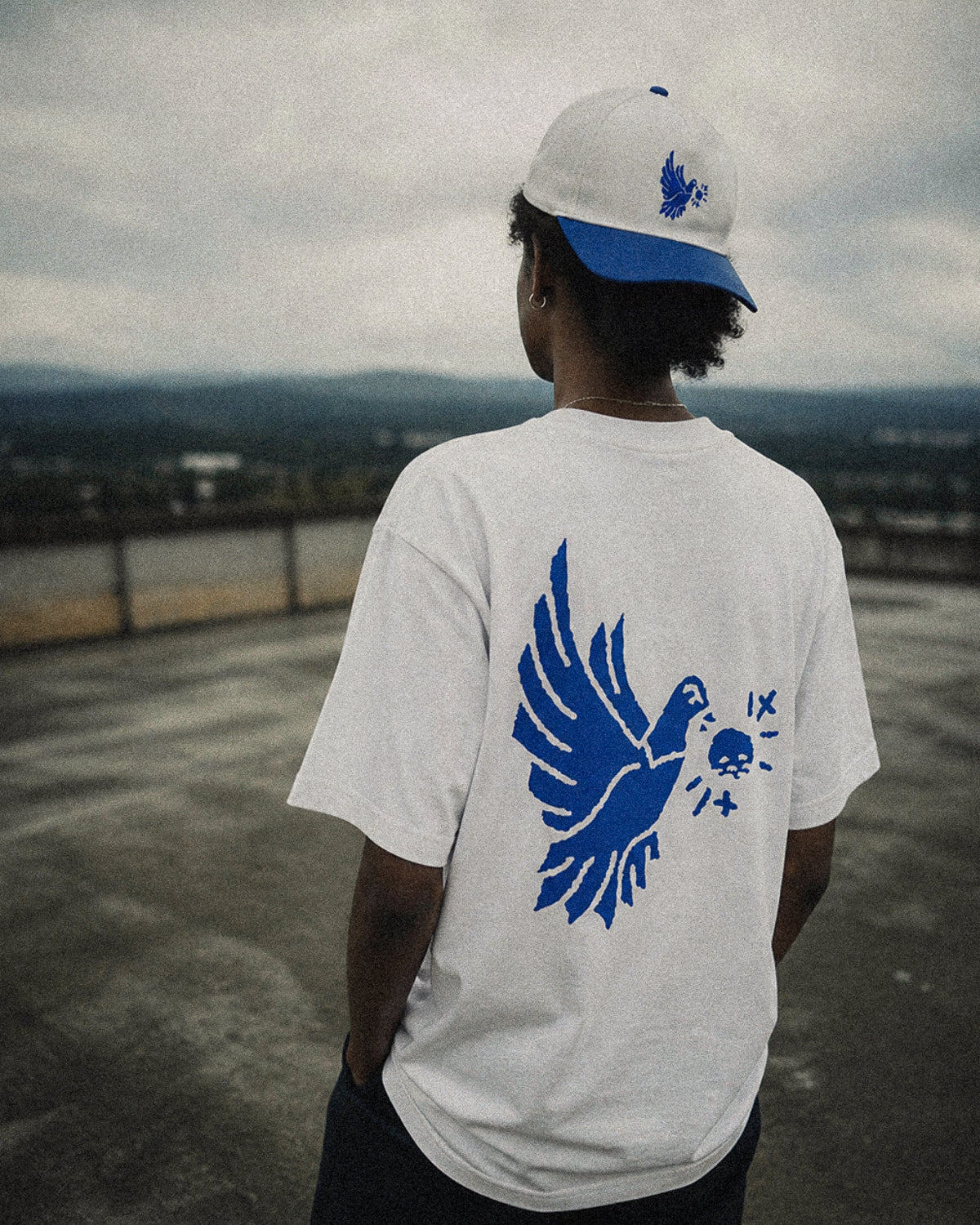 Person wearing a white t-shirt with a blue eagle design on the back, standing on a rooftop with a cityscape in the background.