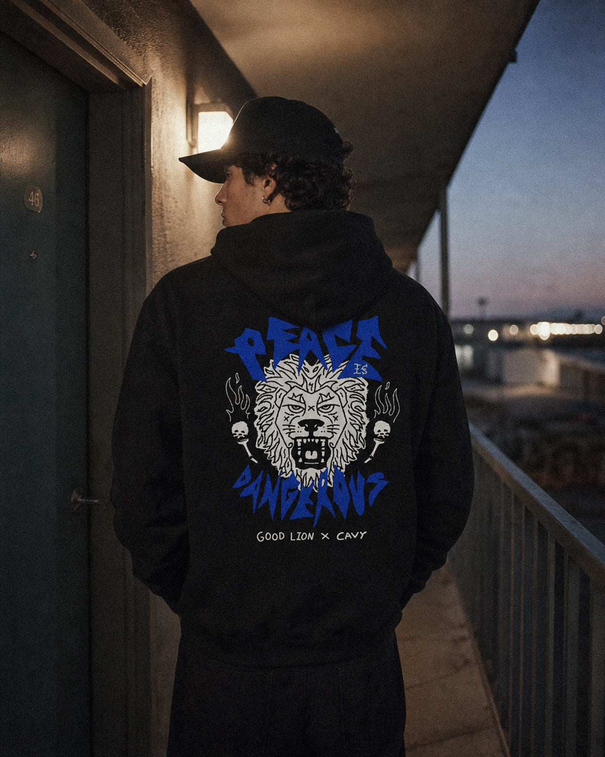 Person wearing a black hoodie with a graphic design on the back, standing on a balcony at dusk.