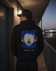 Person wearing a black hoodie with a graphic design on the back, standing on a balcony at dusk.