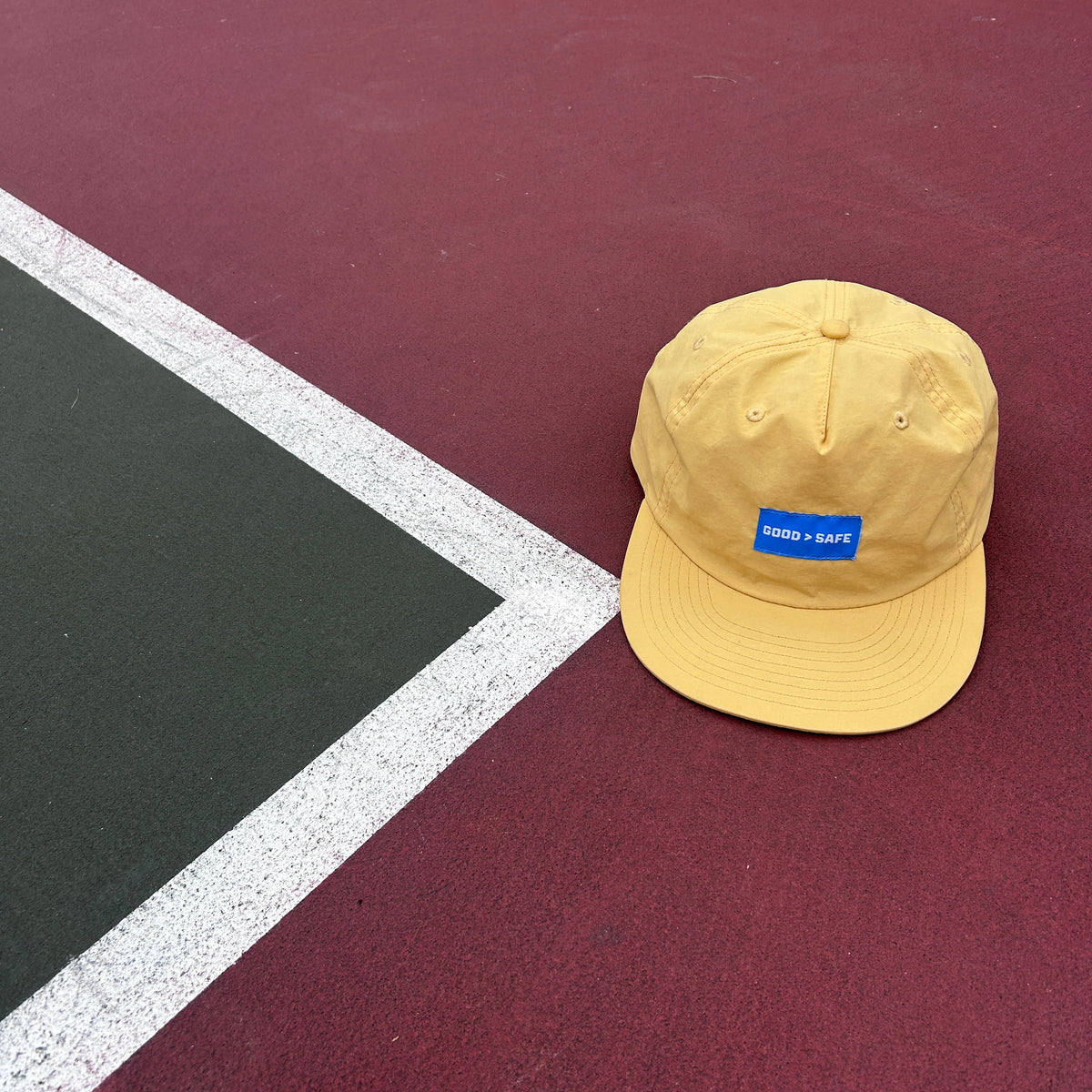 A yellow baseball cap with a blue GOOD > SAFE patch on the front, resting on a red tennis court near the corner line.
