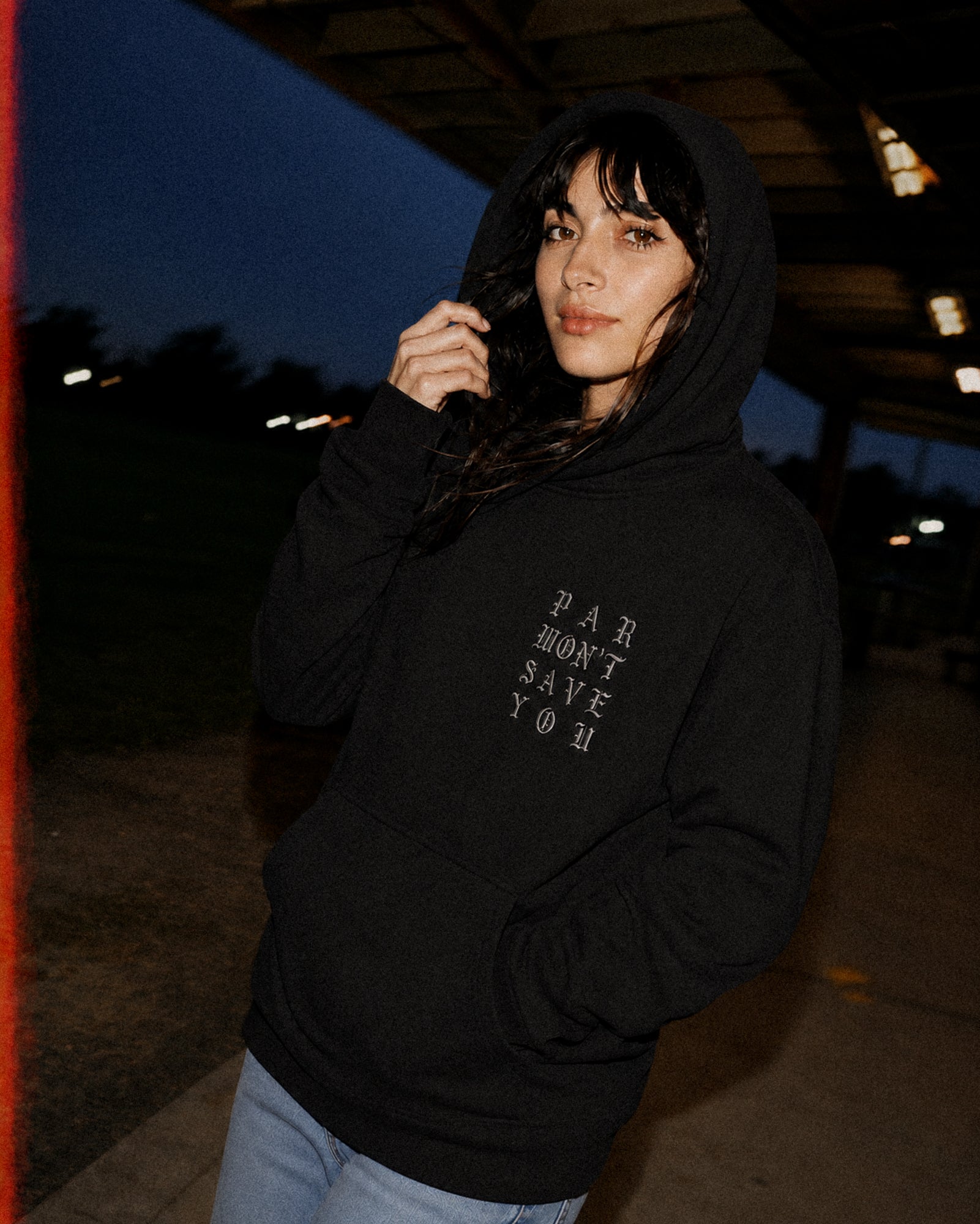 A woman wearing a black pullover hoodie with the text 'PART WON'T SAVE YOU' printed vertically on the left chest, photographed outdoors at dusk under a covered walkway.