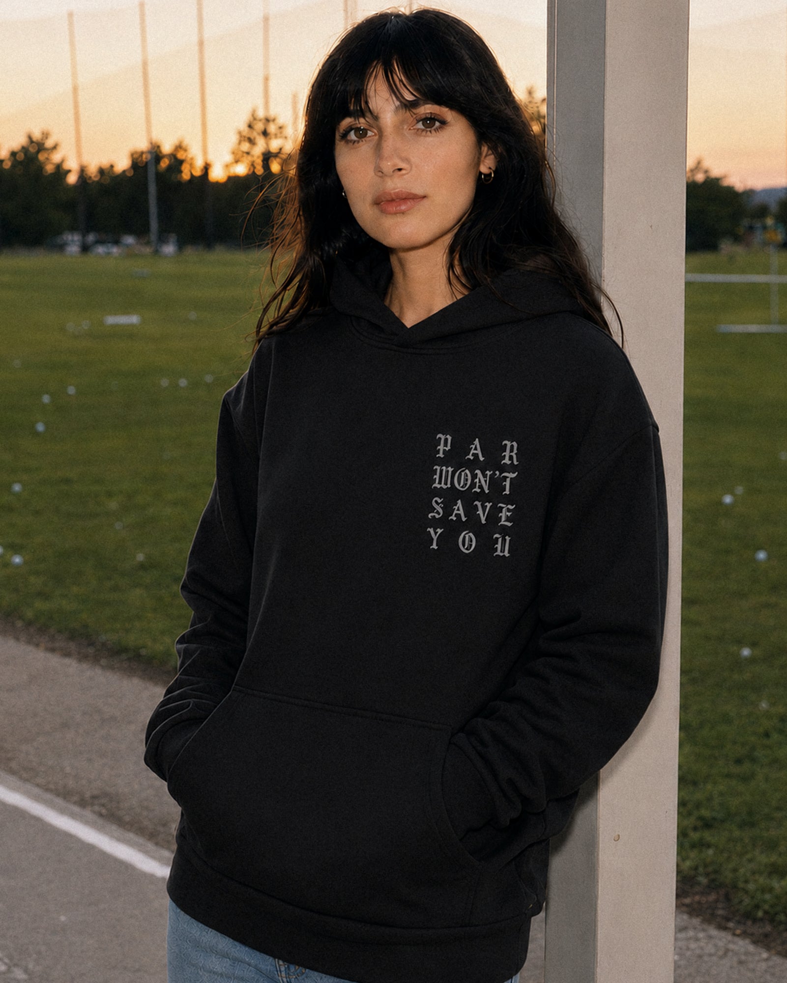 A woman wearing a black pullover hoodie with the text 'PAR WON'T SAVE YOU' printed in white on the left chest, standing outdoors next to a concrete pole at sunset.