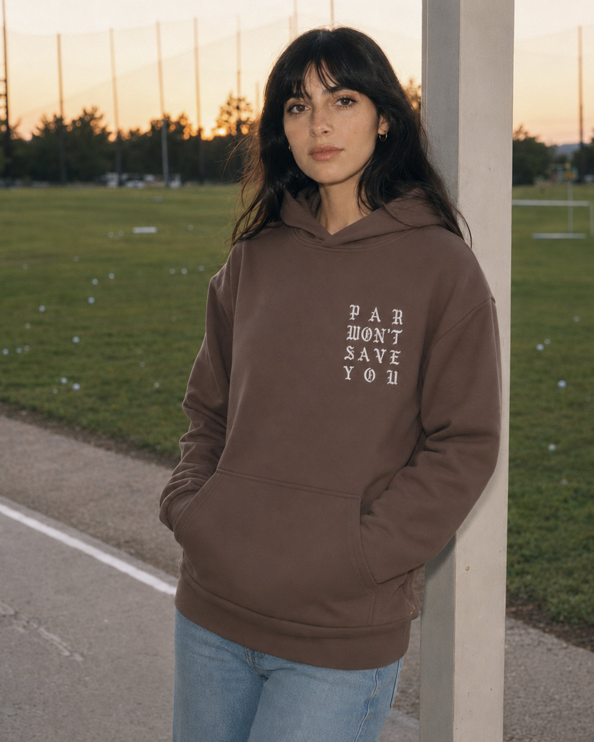 A woman with dark hair and bangs stands outdoors on a track or field path at sunset, wearing a brown pullover hoodie with the text 'PAR DON'T SAVE YOU' printed in white on the chest, paired with light blue jeans.