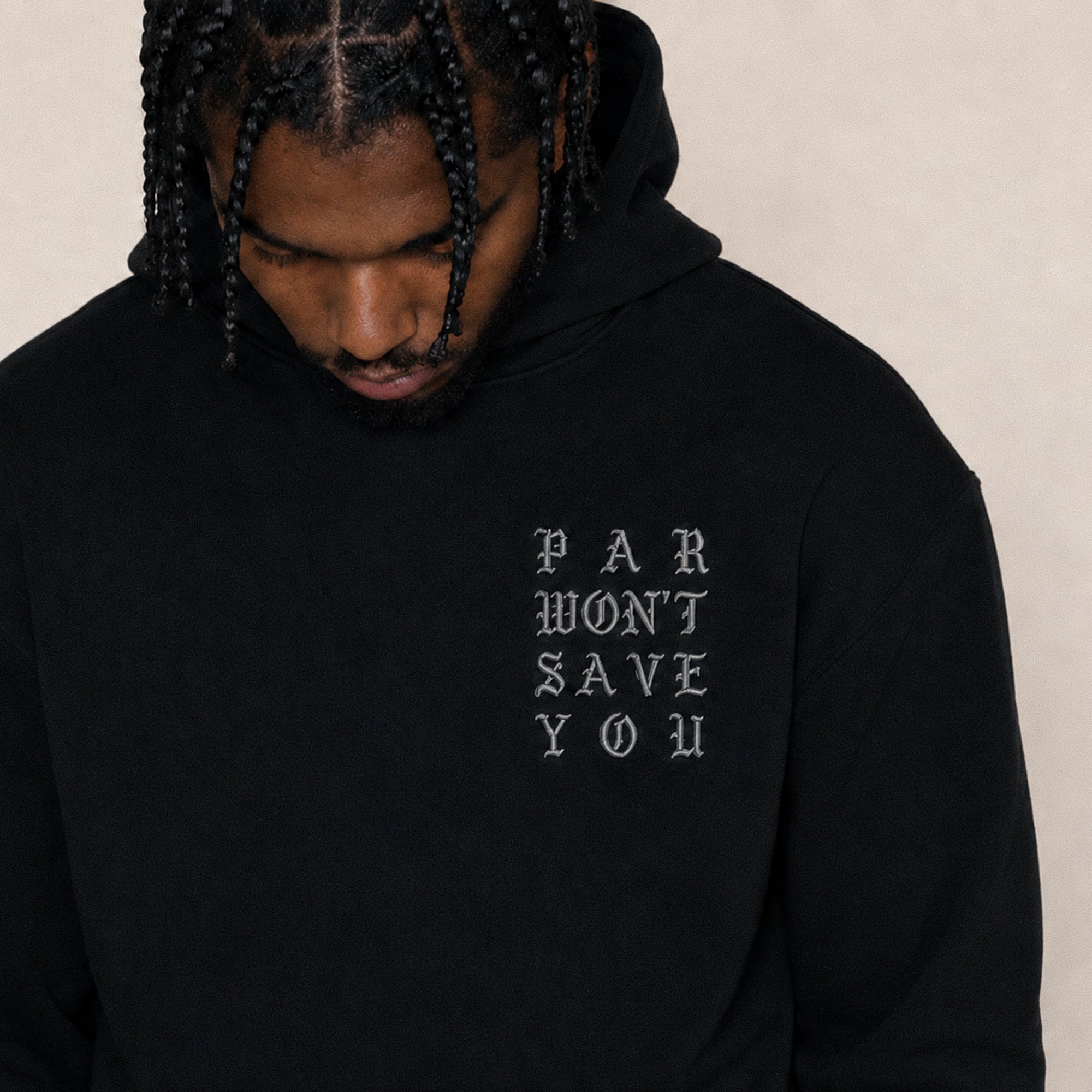 A man wearing a black pullover hoodie with the text 'PAR WON'T SAVE YOU' printed in a light gray gothic font on the left chest, set against a plain beige background.