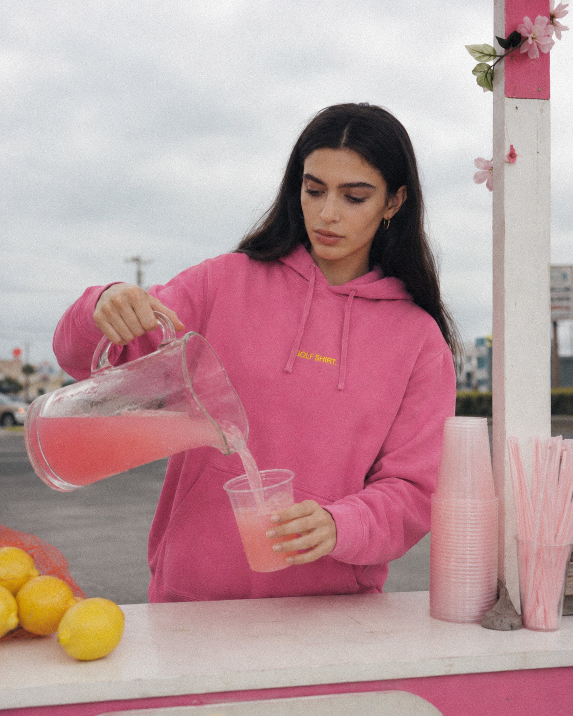 Woman in pink hoodie pouring pink lemonade into a cup at an outdoor stand.