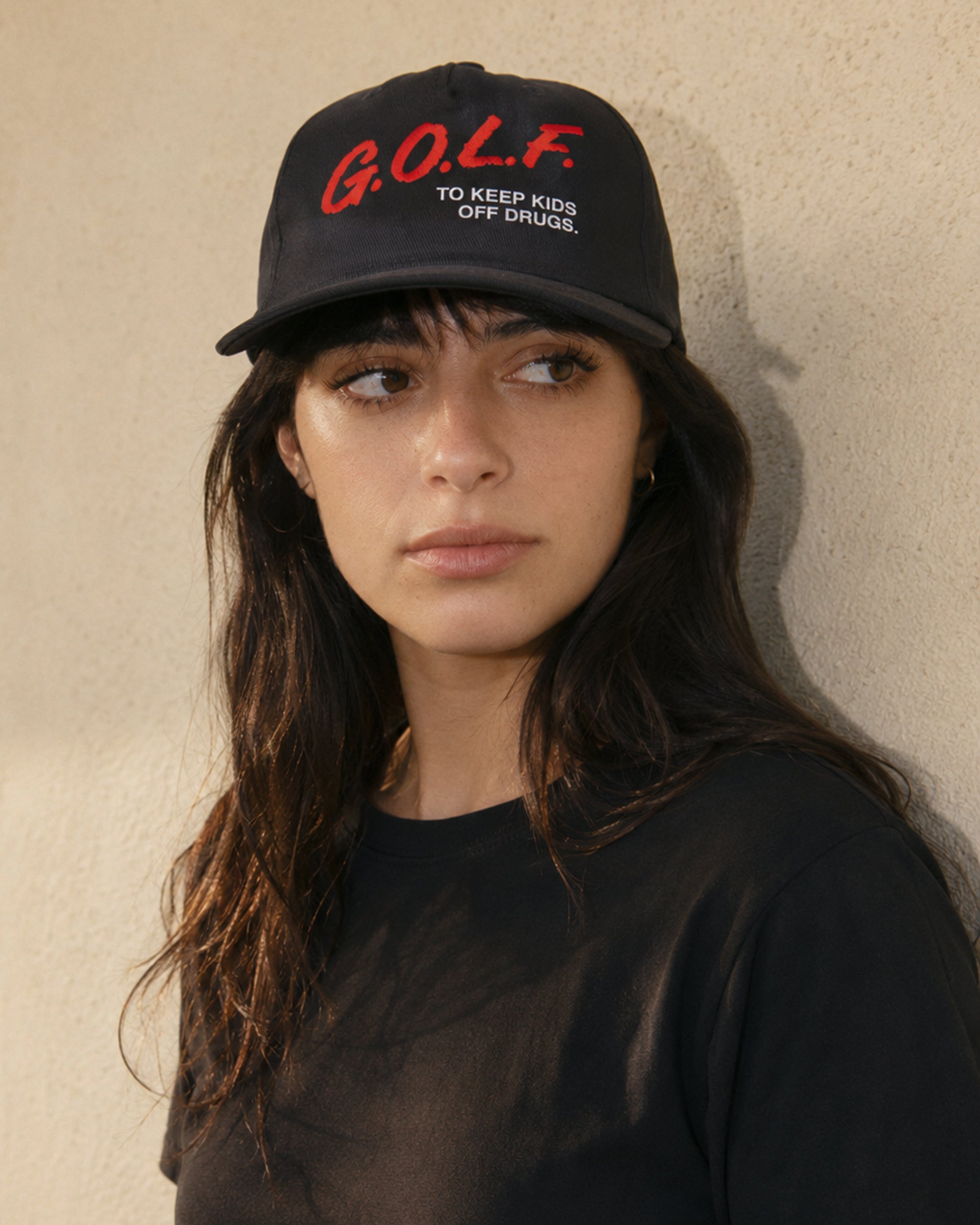 A woman wearing a black baseball cap with red and white embroidered text that reads 'G.O.L.F.