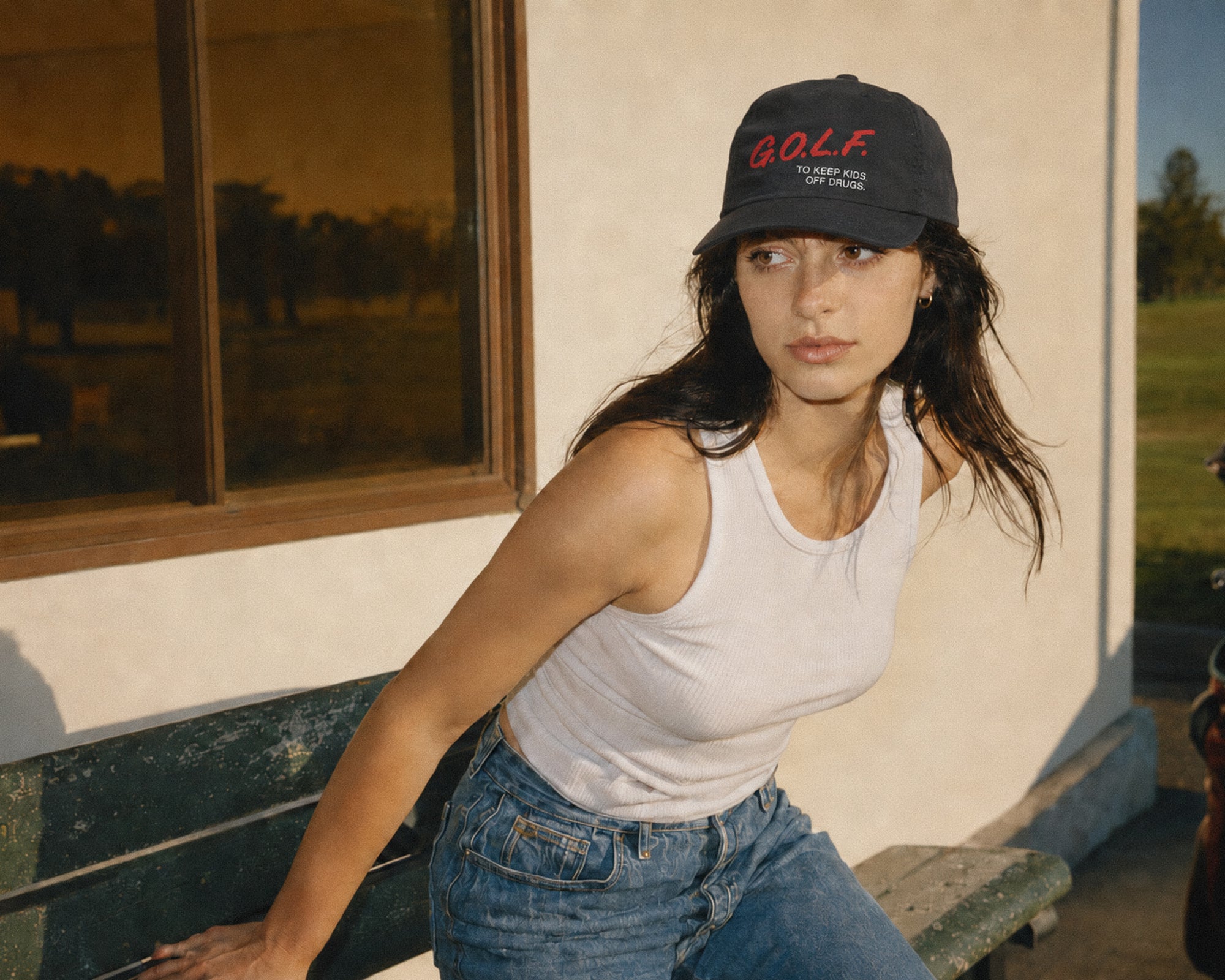 A woman wearing a black baseball cap with red 'GOLF' lettering and a white tank top leans on a bench outdoors next to a building.