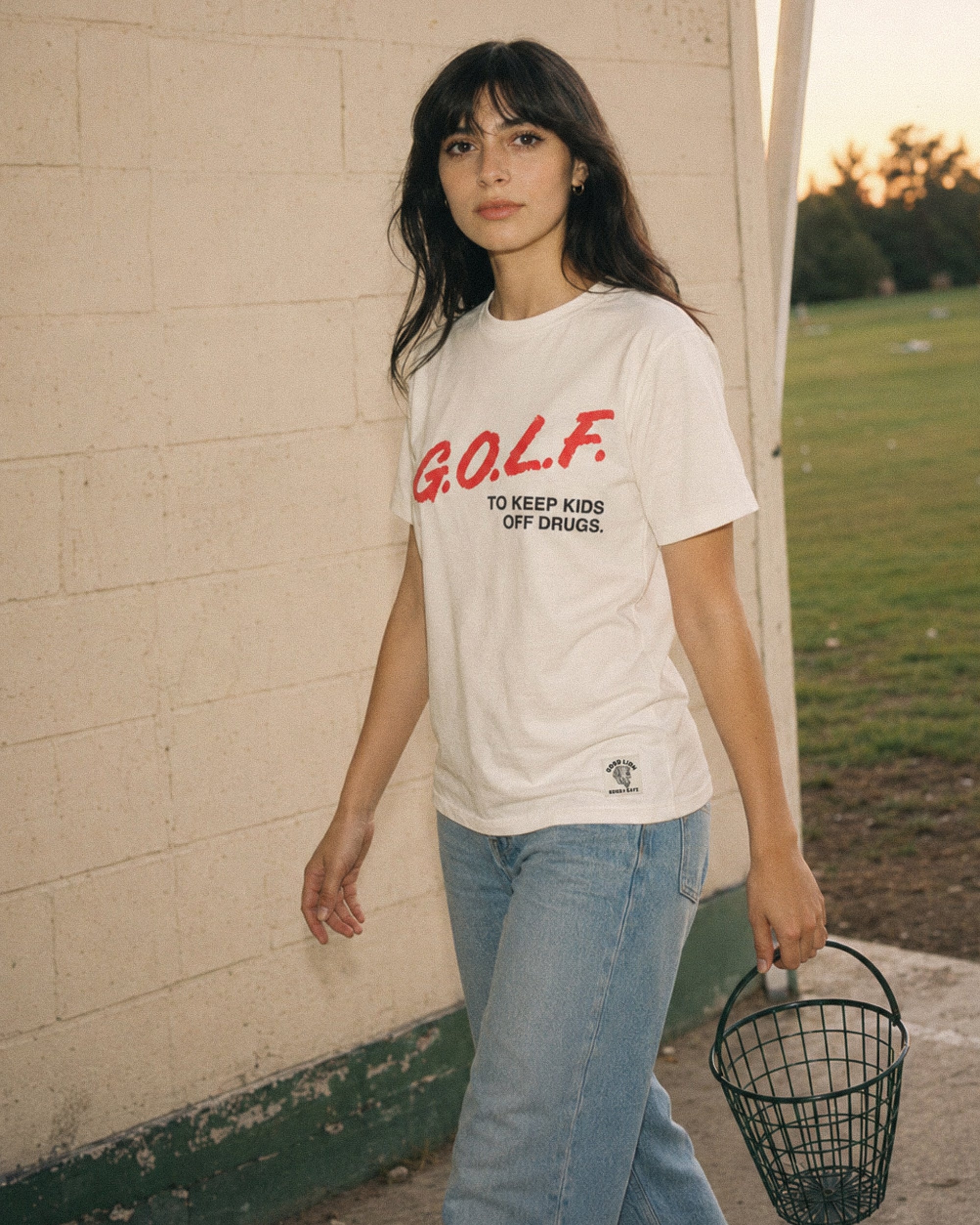 A woman stands outdoors against a light brick wall wearing a white graphic T-shirt with red and black text reading 'GOLF. TO KEEP KIDS OFF DRUGS.