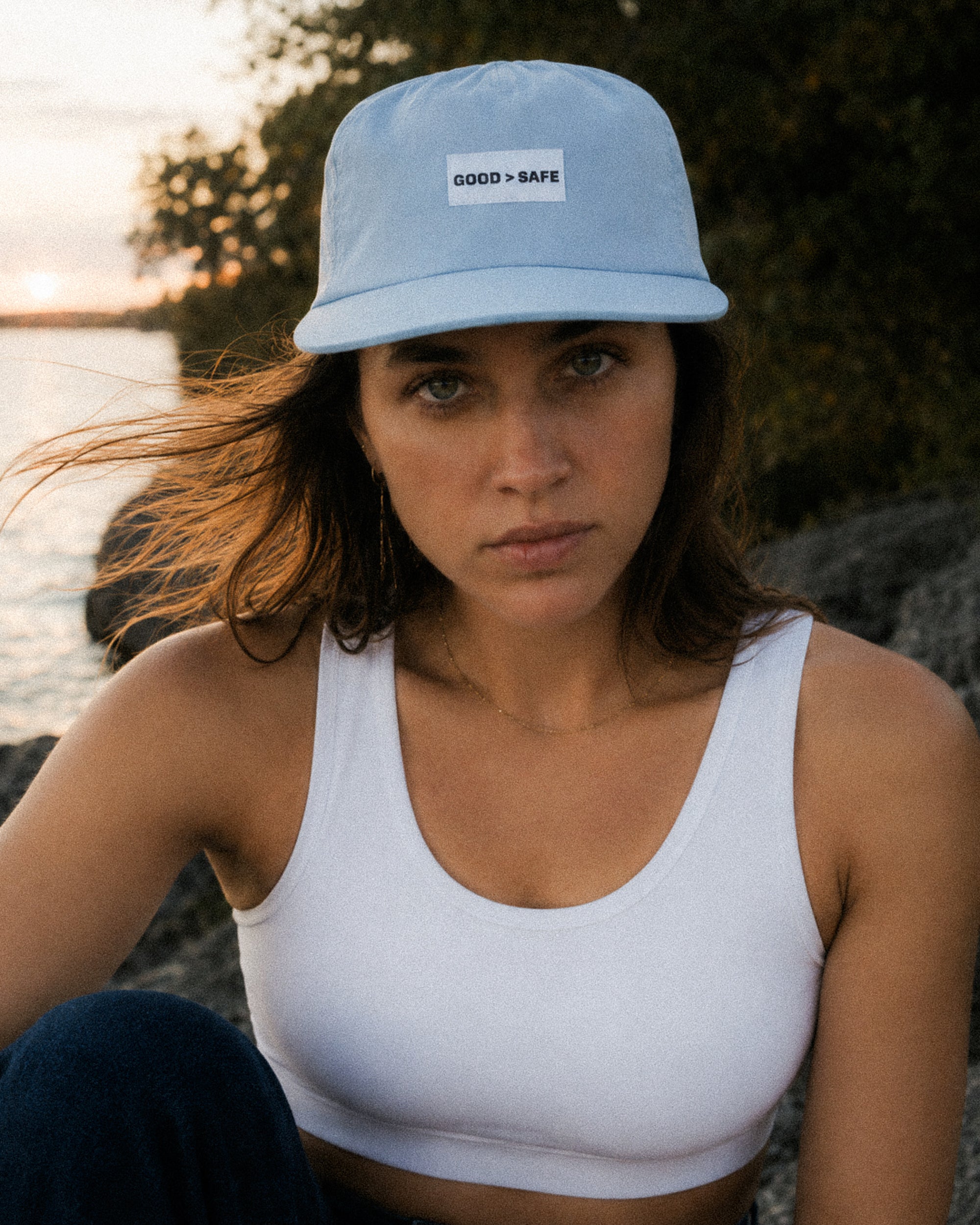 Woman wearing a light blue bucket hat with 'GOOD > SAFE' text, sitting by a body of water.