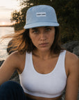 Woman wearing a blue cap with 'GOOD > SAFE' logo by a body of water at sunset.