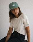 Woman wearing a green cap and white t-shirt with visible branding against a plain background