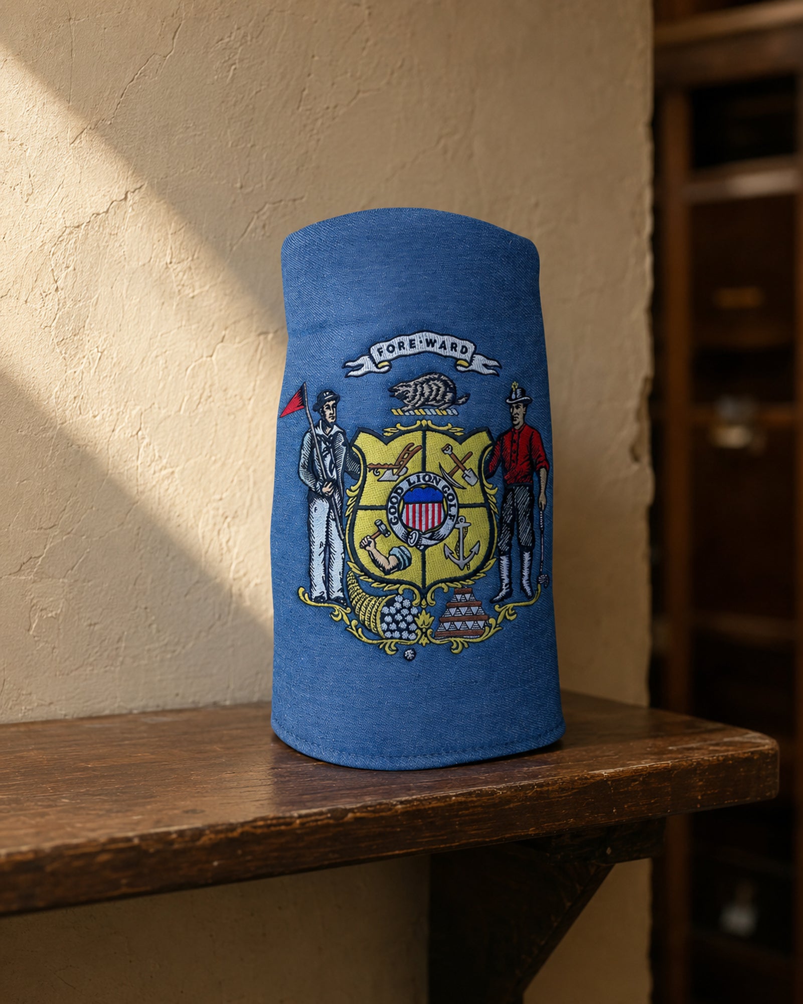 A blue cylindrical denim driver cover with an embroidered Wisconsin state coat of arms design, displayed on a wooden shelf against a textured beige wall.
