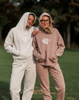 Two people wearing matching hoodie and pants sets standing on grass with trees in the background.