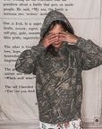 Person wearing a camouflage hoodie with text on it, standing against a wooden wall.